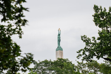 Freedom monument in riga latvia seen through green trees on cloudy day showing national landmark...