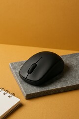 A black wireless mouse on a small stone platform, next to a small notepad with spiral binding, against a yellow-orange background. The mouse appears to be in good condition and ready to use