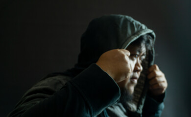 Indonesian man in his 30s with beard and worn gray fabric jacket, hood up and face hidden, moody...