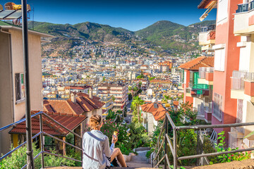 Woman sitting on stairs eating street food, overlooking colorful Mediterranean town and mountains at sunset