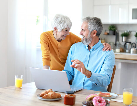 Portrait of an elderly senior couple having breakfast and looking at a laptop at home. Happy healthy affectionate senior couple eating and sitting at kitchen table having fun enjoying morning meal tog