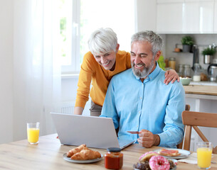Portrait of an elderly senior couple having breakfast and looking at a laptop using a credit card for online shopping at home. Happy healthy affectionate senior couple eating and sitting at kitchen ta