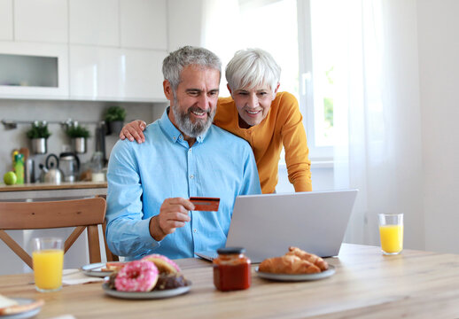 Portrait of an elderly senior couple having breakfast and looking at a laptop using a credit card for online shopping at home. Happy healthy affectionate senior couple eating and sitting at kitchen ta