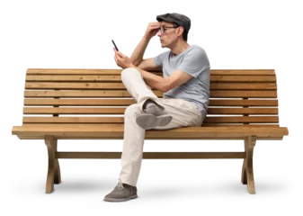 Mature man in his 50s sitting on a wooden bench and using a smartphone, studio shot isolated on white background. Casual clothing, wearing reading glasses and a hat © amedeoemaja