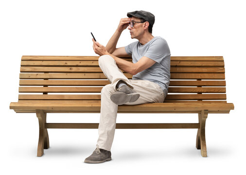 Mature man in his 50s sitting on a wooden bench and using a smartphone, studio shot isolated on white background. Casual clothing, wearing reading glasses and a hat - Powered by Adobe