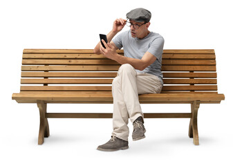Mature man in his 50s sitting on a wooden bench and using a smartphone, studio shot isolated on white background. Casual clothing, wearing reading glasses and a hat