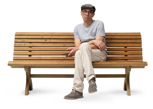 Bored mature man in his 50s sitting on a wooden bench and using a smartphone while looking at the camera. Studio shot, isolated on white background. Casual clothing, wearing reading glasses and a hat