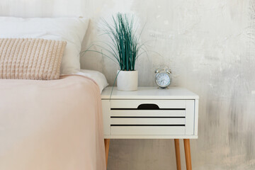 Morning home routine. Houseplant in pot and alarm clock on bedside table in white bedroom interior. Copyspace for text. Minimalist concept. Modern design interior skandinavian bedroom in simple style.