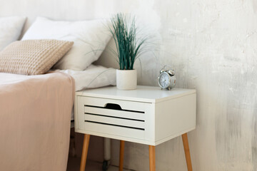Morning home routine. Houseplant in pot and alarm clock on bedside table in white bedroom interior....