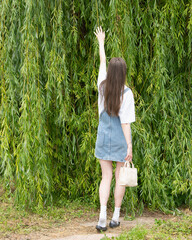 A young woman wearing a denim dress and white shirt stands under a willow tree, reaching up to...