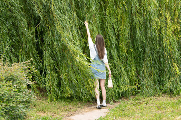 A young woman in a casual outfit reaches out towards the branches of willow trees in a tranquil...