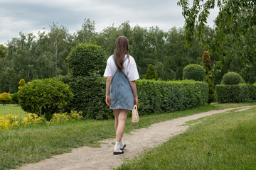 A young woman strolls on a dirt path in a park, dressed casually in a denim dress and holding a...