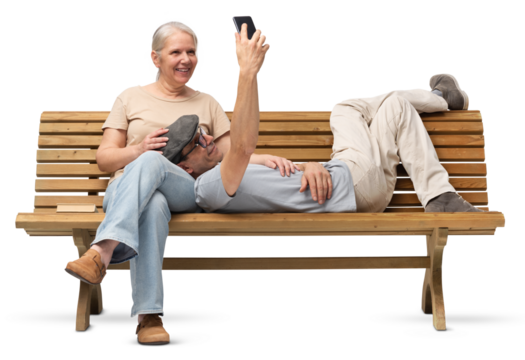 Happy mature couple sitting on a wooden bench. The man is lying down with his head on the woman’s lap while using a smartphone, and she smiles affectionately. Studio shot isolated on white background.