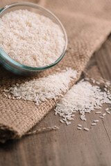 White Rice in Glass Bowl with Grains Spilled on Burlap Fabric, Close-Up of White Rice in a Glass Bowl on Rustic Sackcloth, Glass Bowl Full of Rice on Wooden Table, Organic White Rice with Glass Bowl


