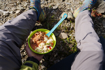 Tourist lunch served in a silicone camping bowl placed on the ground between the traveler's legs. Outdoor meal during a hiking trip in nature.