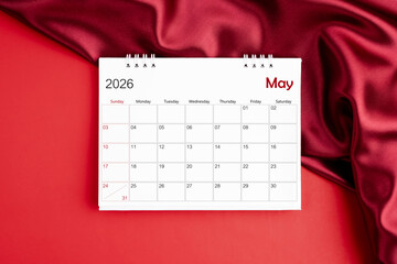 Top view of May 2026 desk calendar on red silk color background.