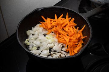 Home cooking. On the stove in a cast iron frying pan onions and carrots are fried in a hot frying...