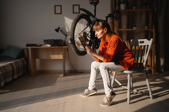 Middle aged woman repairing a broken bicycle at home. Female cyclist inspecting bicycle with digital tablet.