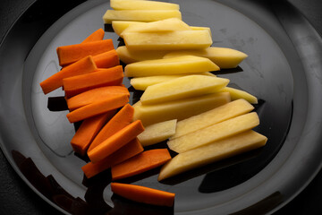 Raw potatoes cut into large strips and raw carrots cut into strips on a black plate, top view, side...
