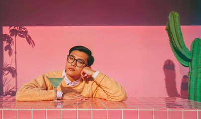 Young Asian nerd sits at a checkered table with blue cocktail, wearing vintage retro outfit with a pastel pink wall in the background.  Back to school conceptual copy space