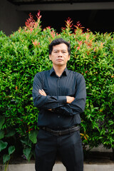 Confident asian man with arms crossed stands before vibrant green foliage and red tips in daylight