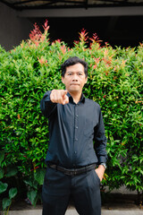 Asian man in formal attire pointing forward, green shrubbery background, selective focus and