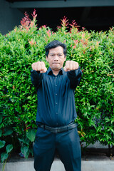 Middle-aged man in black outfit poses with fists clenched in front of lush green foliage background