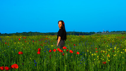 A young woman with long dark hair sitting on a green meadow in summer, surrounded by tall grass and wildflowers, enjoying the warm golden sunlight.