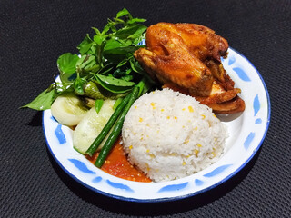 Lalapan ayam goreng. Crispy golden fried chicken dishes, with bright red chili sauce, fresh cucumber slices and a few basil leaves on a white enamel plate decorated with blue.