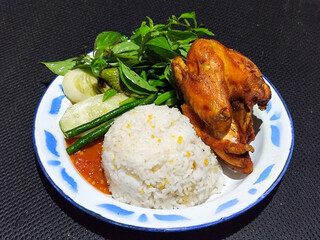 Lalapan ayam goreng. Crispy golden fried chicken dishes, with bright red chili sauce, fresh cucumber slices and a few basil leaves on a white enamel plate decorated with blue.