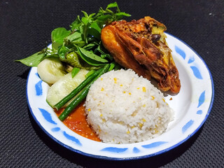 Lalapan ayam goreng. Crispy golden fried chicken dishes, with bright red chili sauce, fresh cucumber slices and a few basil leaves on a white enamel plate decorated with blue.