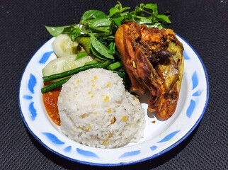 Lalapan ayam goreng. Crispy golden fried chicken dishes, with bright red chili sauce, fresh cucumber slices and a few basil leaves on a white enamel plate decorated with blue.