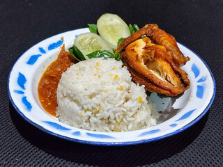 Lalapan ayam goreng. Crispy golden fried chicken dishes, with bright red chili sauce, fresh cucumber slices and a few basil leaves on a white enamel plate decorated with blue.