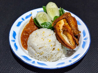 Lalapan ayam goreng. Crispy golden fried chicken dishes, with bright red chili sauce, fresh cucumber slices and a few basil leaves on a white enamel plate decorated with blue.