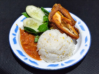 Lalapan ayam goreng. Crispy golden fried chicken dishes, with bright red chili sauce, fresh cucumber slices and a few basil leaves on a white enamel plate decorated with blue.