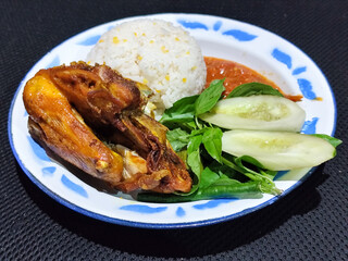 Lalapan ayam goreng. Crispy golden fried chicken dishes, with bright red chili sauce, fresh cucumber slices and a few basil leaves on a white enamel plate decorated with blue.