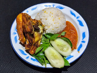 Lalapan ayam goreng. Crispy golden fried chicken dishes, with bright red chili sauce, fresh cucumber slices and a few basil leaves on a white enamel plate decorated with blue.