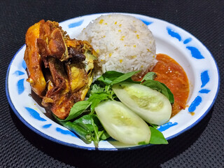 Lalapan ayam goreng. Crispy golden fried chicken dishes, with bright red chili sauce, fresh cucumber slices and a few basil leaves on a white enamel plate decorated with blue.