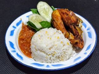 Lalapan ayam goreng. Crispy golden fried chicken dishes, with bright red chili sauce, fresh cucumber slices and a few basil leaves on a white enamel plate decorated with blue.