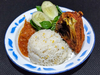 Lalapan ayam goreng. Crispy golden fried chicken dishes, with bright red chili sauce, fresh cucumber slices and a few basil leaves on a white enamel plate decorated with blue.