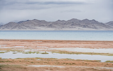 Grass and stone shore of Lake Karakul with blue water and mountain peaks in the background, cloudy...