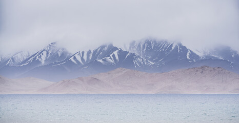 Grass and stone shore of Lake Karakul with blue water and mountain peaks in the background, cloudy...
