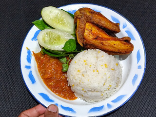 Lalapan ayam goreng. Crispy golden fried chicken dishes, with bright red chili sauce, fresh cucumber slices and a few basil leaves on a white enamel plate decorated with blue.