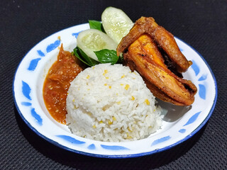Lalapan ayam goreng. Crispy golden fried chicken dishes, with bright red chili sauce, fresh cucumber slices and a few basil leaves on a white enamel plate decorated with blue.