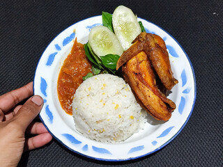 Lalapan ayam goreng. Crispy golden fried chicken dishes, with bright red chili sauce, fresh cucumber slices and a few basil leaves on a white enamel plate decorated with blue.