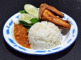 Lalapan ayam goreng. Crispy golden fried chicken dishes, with bright red chili sauce, fresh cucumber slices and a few basil leaves on a white enamel plate decorated with blue.