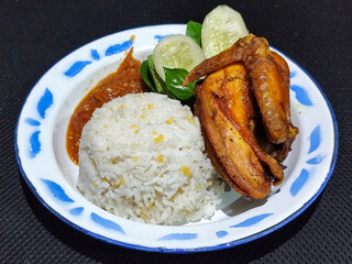 Lalapan ayam goreng. Crispy golden fried chicken dishes, with bright red chili sauce, fresh cucumber slices and a few basil leaves on a white enamel plate decorated with blue.
