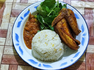 Lalapan ayam goreng. Crispy golden fried chicken dishes, with bright red chili sauce, fresh cucumber slices and a few basil leaves on a white enamel plate decorated with blue.