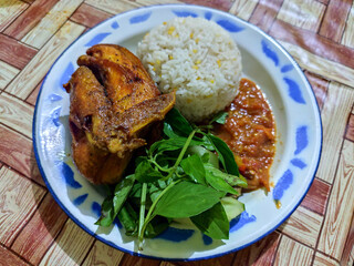 Lalapan ayam goreng. Crispy golden fried chicken dishes, with bright red chili sauce, fresh cucumber slices and a few basil leaves on a white enamel plate decorated with blue.