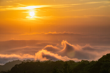 A spectacular golden sunrise illuminates dramatic, wavy clouds rolling over the mountains of Shulin District New Taipei City Taiwan, with faint city silhouettes below.
